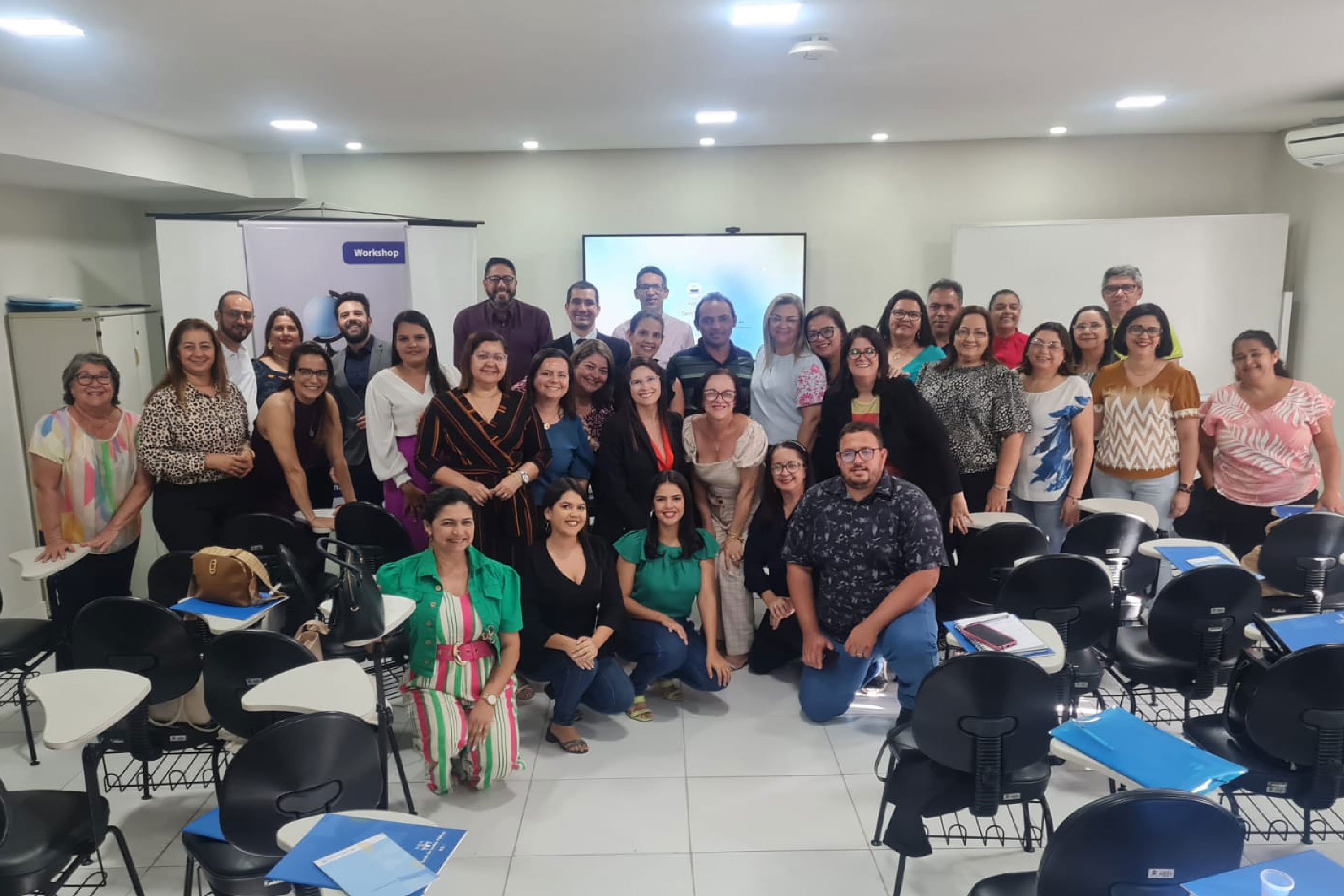 Grupo de pessoas posando para foto, numa sala de aula. Atrás das pessoas, fundo branco com presença de painéis de recursos audiovisuais.
