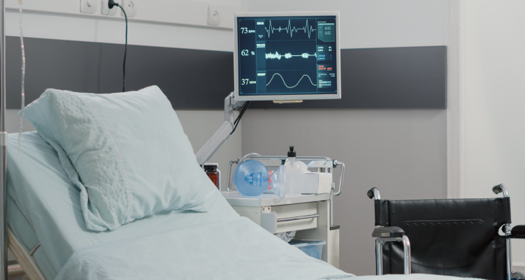 Foto mostra um leito hospitalar com um monitor instalado na parede. No canto direito da foto, há uma cadeira de rodas.
